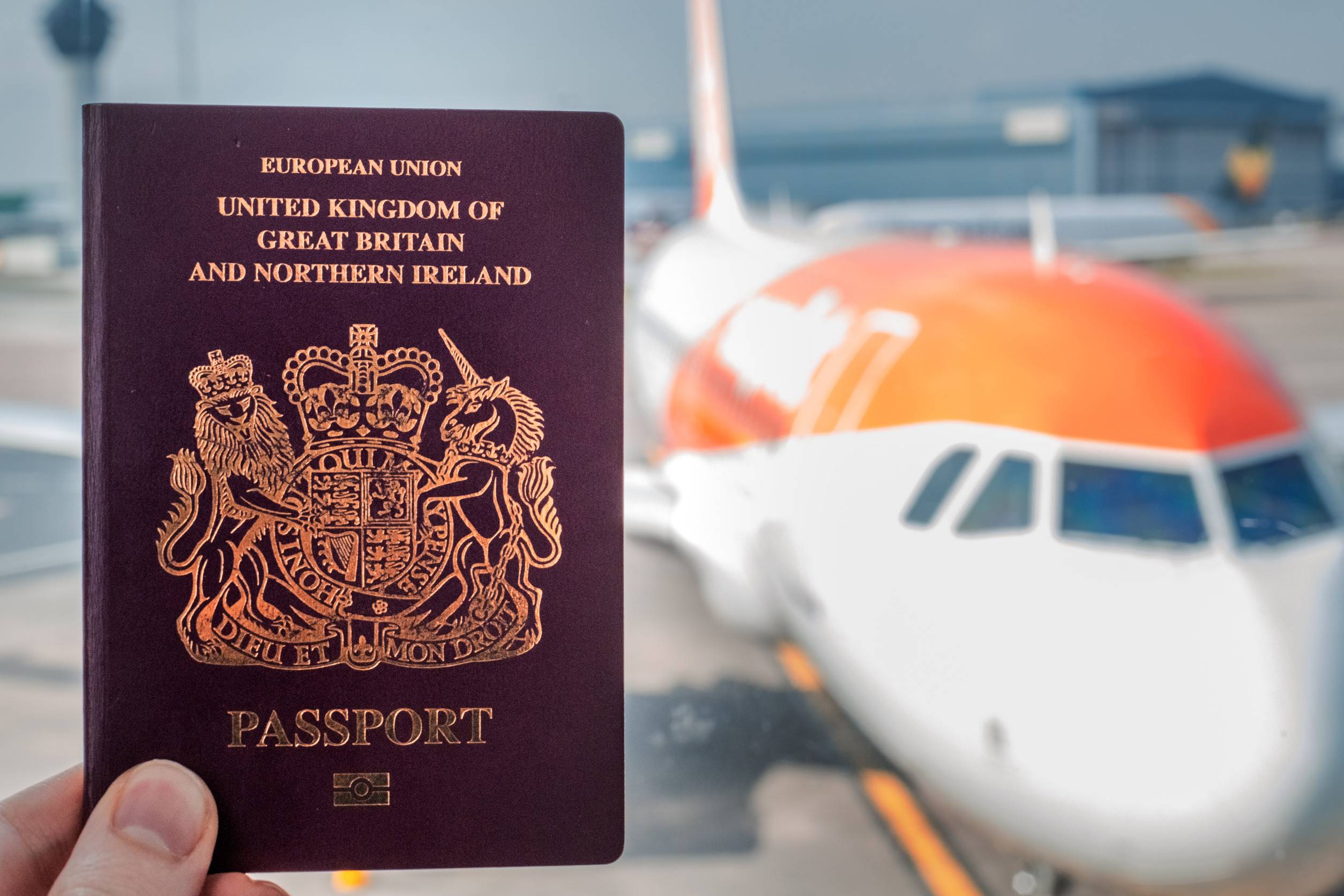 Red British Passport Held Up Against A Background Of A Generic Plane On A Brigh